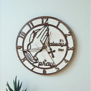 May include: Wooden wall clock with a light wood face and a dark brown frame. The clock features Roman numerals and hands, with the words "It's always time to pray" and an image of praying hands.