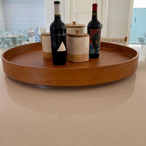May include: A round, wooden serving tray holds two wine bottles and two decorative canisters. One wine bottle has a white triangle on the label, the other has a colorful portrait. The canisters are white and beige with wooden lids.