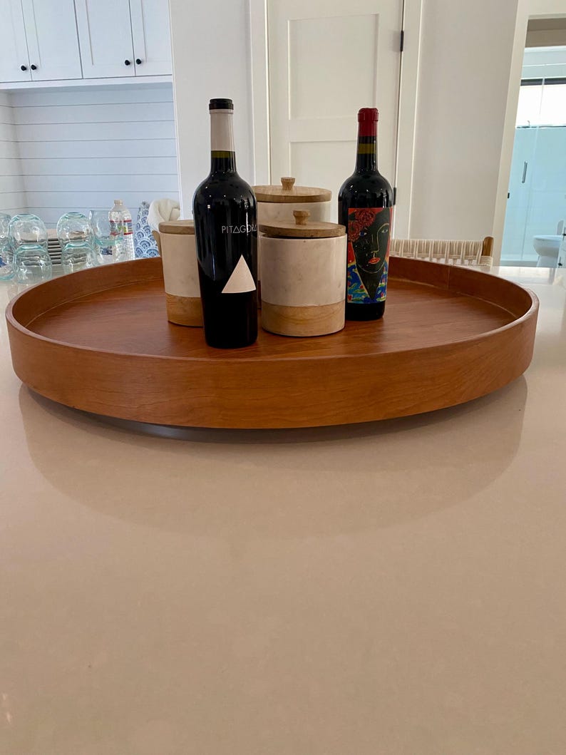 May include: A round, wooden serving tray holds two wine bottles and two decorative canisters. One wine bottle has a white triangle on the label, the other has a colorful portrait. The canisters are white and beige with wooden lids.