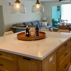 May include: A kitchen island with a light-colored countertop and wooden cabinets. A wooden tray holds bottles and jars. Two glass pendant lights hang above the island. A living room with a blue sofa is visible in the background.