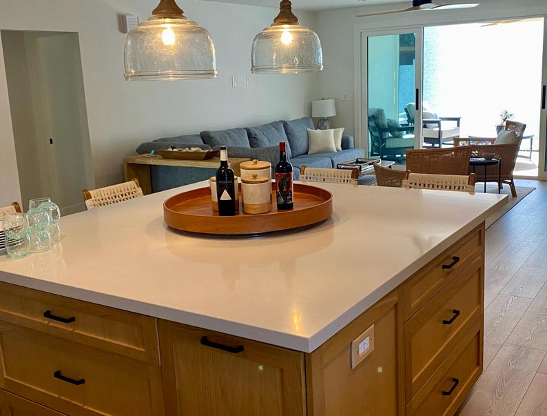 May include: A kitchen island with a light-colored countertop and wooden cabinets. A wooden tray holds bottles and jars. Two glass pendant lights hang above the island. A living room with a blue sofa is visible in the background.