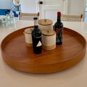May include: A round, wooden serving tray holds two wine bottles and two small canisters with wooden lids. One wine bottle has a white label with the word "PITAGOR" and a triangle. The other bottle has a colorful label.