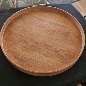 May include: A round wooden tray with a raised edge. The tray is made of light brown wood and has a natural wood grain pattern. The tray is sitting on a black surface.