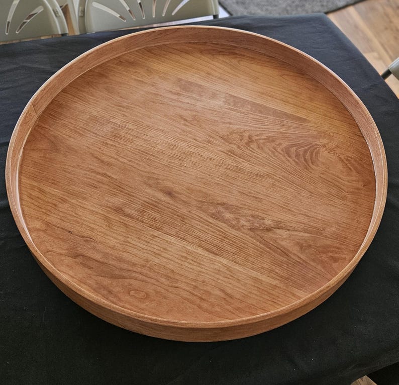 May include: A round wooden tray with a raised edge. The tray is made of light brown wood and has a natural wood grain pattern. The tray is sitting on a black surface.