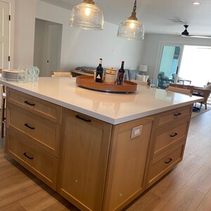 May include: A light wood kitchen island with a white countertop. The island has drawers and cabinets with black handles. Two glass pendant lights hang above the island. A wooden tray holds wine bottles and jars.
