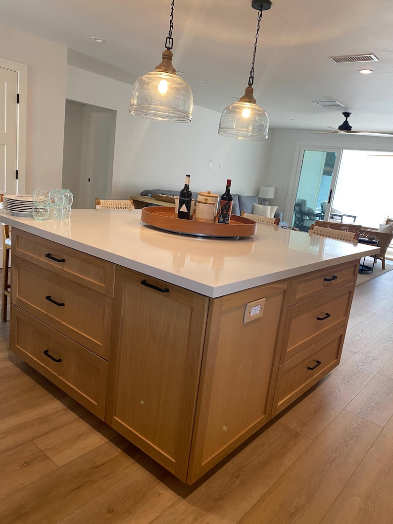 May include: A light wood kitchen island with a white countertop. The island has drawers and cabinets with black handles. Two glass pendant lights hang above the island. A wooden tray holds wine bottles and jars.