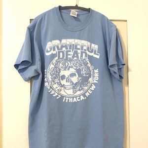 May include: A light blue t-shirt with a white graphic of a skull with roses and the text "Grateful Dead" and "May 8th 1977 Ithaca, New York".