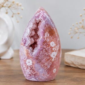 Puede incluir: Una geoda de ágata rosa pulida en forma de lágrima que presenta una cavidad central llena de pequeñas estructuras cristalinas brillantes. La superficie exterior muestra un acabado liso y translúcido con inclusiones blancas en forma de flor.