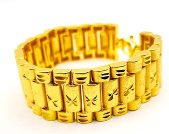 Puede incluir: Un brazalete dorado con un diseño tipo reloj. El brazalete presenta eslabones rectangulares con recortes en forma de estrella. El cierre es visible en el lado derecho. El brazalete está hecho de un material metálico brillante.