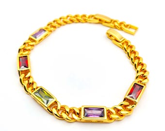 Puede incluir: Pulsera de cadena dorada con piedras preciosas rectangulares de colores. La pulsera presenta una serie de eslabones dorados, cada uno con una piedra preciosa de diferente color: rojo, verde, morado y amarillo. La pulsera se muestra sobre un fondo blanco.