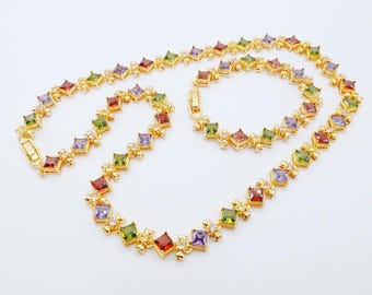 Può includere: Un set di collana e bracciale color oro. I gioielli presentano pietre preziose multicolori a taglio quadrato in rosso, verde, viola e giallo. Le pietre sono incastonate in un motivo ripetuto, collegate da piccoli anelli color oro.