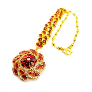 May include: A gold-toned necklace with a large, ornate pendant. The pendant features a central cluster of red gemstones surrounded by a spiral of red gems and smaller clear stones. The necklace chain is made of small gold beads.