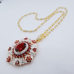 May include: A necklace featuring a large, oval-cut deep red gemstone pendant surrounded by smaller red and clear stones set in a silver-toned metal frame. The pendant hangs from a chain composed of alternating gold-toned and silver-toned cylindrical links.