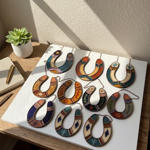 Puede incluir: Una colección de colgantes en forma de herradura con diseños coloridos y variados, incluyendo patrones florales y geométricos. Cada colgante está conectado a una cadena. Los colgantes se exhiben sobre una superficie blanca, con una pequeña planta suculenta en el fondo.