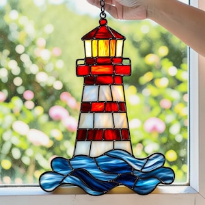 Op de afbeelding: Gebrandschilderde glazen vuurtoren in rood, wit en blauw. De vuurtoren heeft een rode top en een gestreept lichaam. De basis heeft blauwe golven. Een ketting is bevestigd om op te hangen. Decoratief nautisch thema.