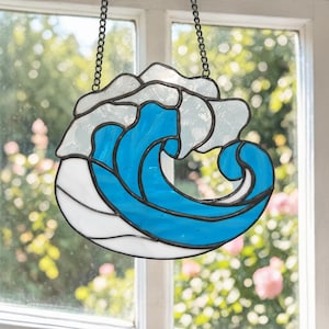 Puede incluir: Pieza de arte de vidriera con forma de ola en diseño azul y blanco. La ola está delineada en negro y cuelga de una cadena. El fondo es una vista borrosa de una ventana y vegetación.