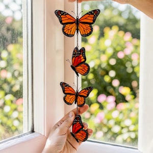 Puede incluir: Un colgante decorativo con cuatro adornos de mariposas naranjas y negras. Las mariposas est&aacute;n ensartadas en una cadena plateada. Las mariposas se sostienen frente a una ventana con un fondo borroso de follaje verde y rosa.