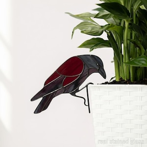 以下が含まれることがあります： 濃い灰色の頭と翼、深紅色の体を持つステンドグラスのカラスの装飾。カラスは、白い織りのプランターの端に掛けるように設計された黒い金属製のブラケットにとまっています。「real stained glass」の文字が見えます。