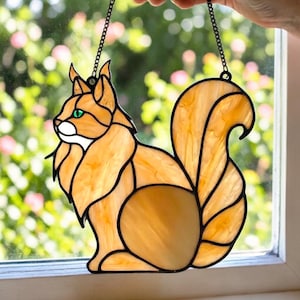May include: A stained glass squirrel suncatcher with a chain for hanging. The squirrel is in shades of orange and yellow with black outlines and green eyes. The suncatcher is displayed in front of a window.