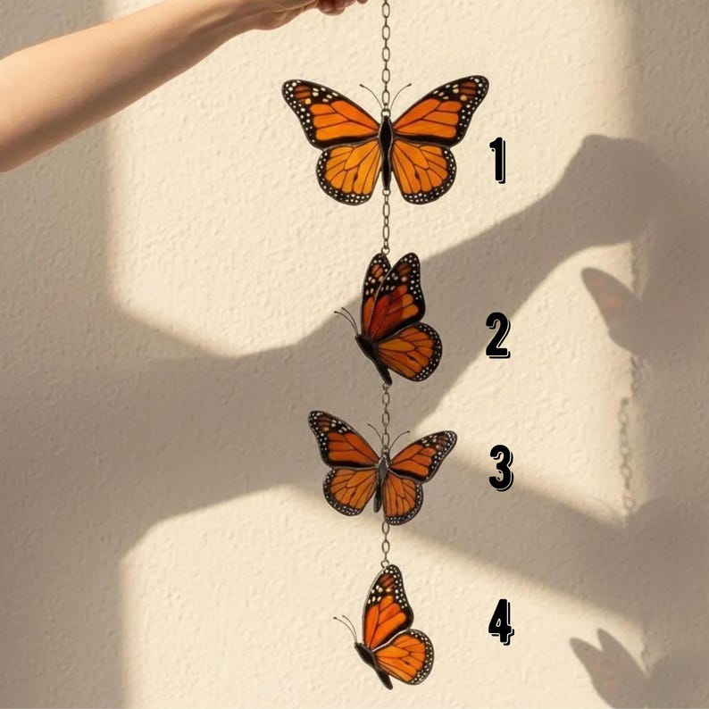 Puede incluir: Colgante decorativo con cuatro adornos de mariposa naranja y negra. Las mariposas est&aacute;n conectadas por una cadena y est&aacute;n suspendidas frente a una pared de color neutro. Las mariposas tienen patrones de alas detallados.
