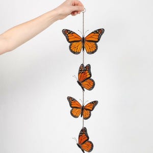 Puede incluir: Un adorno colgante decorativo con cuatro r&eacute;plicas de mariposas monarca de color naranja y negro. Las mariposas est&aacute;n suspendidas de una cadena plateada, y cada mariposa muestra patrones de alas detallados. El adorno es sostenido por una mano sobre un fondo blanco.