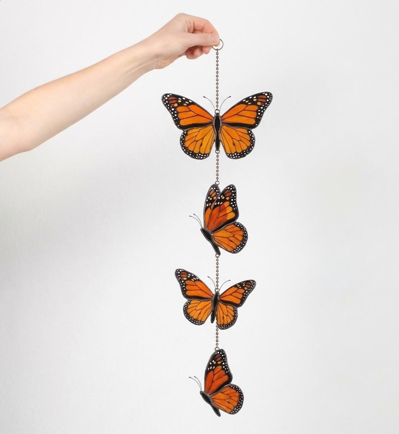 Puede incluir: Un adorno colgante decorativo con cuatro r&eacute;plicas de mariposas monarca de color naranja y negro. Las mariposas est&aacute;n suspendidas de una cadena plateada, y cada mariposa muestra patrones de alas detallados. El adorno es sostenido por una mano sobre un fondo blanco.
