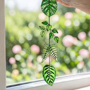 Puede incluir: Móvil de plantas de vidriera con tres diseños de hojas: una gran hoja de Monstera, una enredadera y una hoja de helecho. Las hojas son de varios tonos de verde y están conectadas por una delicada cadena. El fondo es una vista borrosa de un jardín.