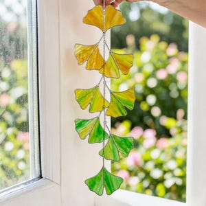 Puede incluir: Móvil de hojas de ginkgo de vidriera con hojas amarillas y verdes. Las hojas están conectadas por una cadena plateada. El móvil cuelga frente a una ventana con un fondo borroso de follaje verde y rosa.