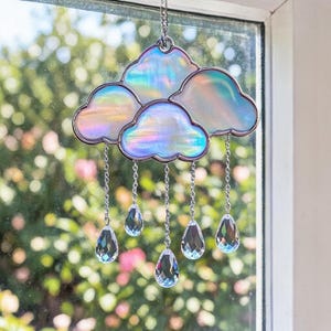Puede incluir: Un atrapasoles decorativo en forma de nube con paneles de vidrio iridiscente y un marco de metal plateado. La nube tiene cinco secciones y cuelga de una cadena. Gotas de cristal transparente cuelgan de la nube.