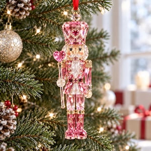 May include: A faceted pink crystal nutcracker ornament featuring a tall crown with a heart-shaped accent, a matching heart-shaped staff, and gold-tone metal detailing. The figure is suspended by a red ribbon and displays clear and pink crystal components.
