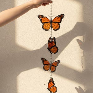 Puede incluir: M&oacute;vil decorativo colgante con cuatro adornos de mariposas monarca naranjas y negras. Las mariposas est&aacute;n conectadas por una delicada cadena y suspendidas sobre un fondo neutro. El m&oacute;vil est&aacute; sostenido por una mano.