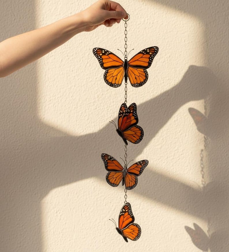 Puede incluir: M&oacute;vil decorativo colgante con cuatro adornos de mariposas monarca naranjas y negras. Las mariposas est&aacute;n conectadas por una delicada cadena y suspendidas sobre un fondo neutro. El m&oacute;vil est&aacute; sostenido por una mano.