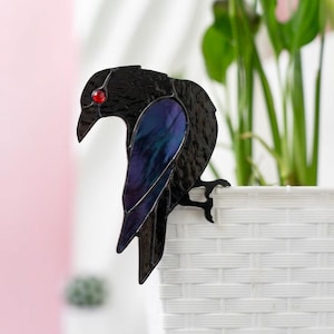 May include: A black stained glass raven with a red eye and iridescent blue and purple wing detail. The raven is perched on the edge of a white woven planter, with a green plant in the background. Decorative garden art.