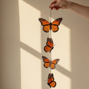 Puede incluir: M&oacute;vil decorativo con cuatro adornos de mariposas monarca naranjas y negras suspendidos por una cadena plateada. Las mariposas est&aacute;n detalladas con contornos negros y puntos blancos, creando un acento inspirado en la naturaleza.