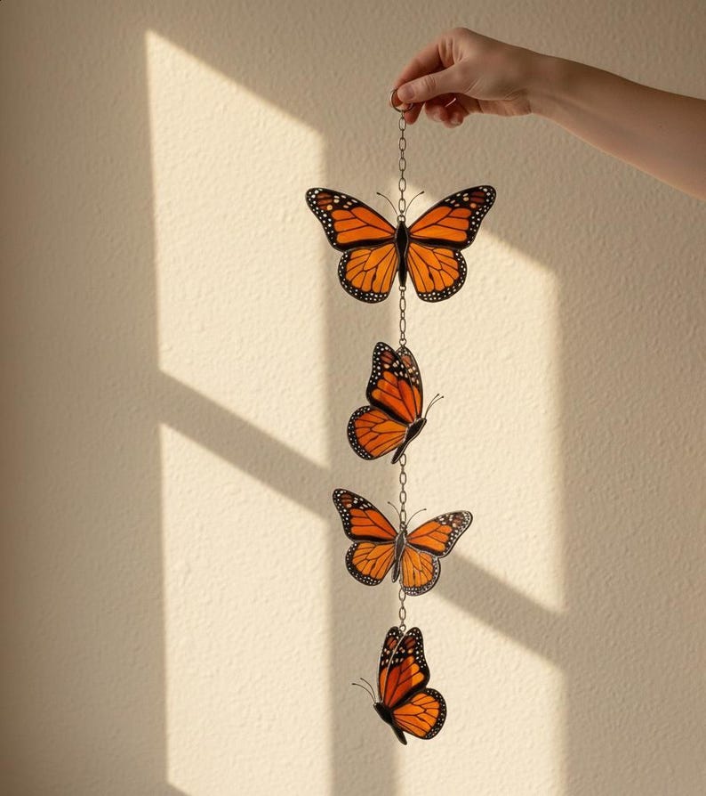 Puede incluir: M&oacute;vil decorativo con cuatro adornos de mariposas monarca naranjas y negras suspendidos por una cadena plateada. Las mariposas est&aacute;n detalladas con contornos negros y puntos blancos, creando un acento inspirado en la naturaleza.