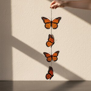 Puede incluir: Colgante decorativo con cuatro adornos de mariposas monarca naranjas y negras suspendidos por una cadena plateada. Las mariposas son sostenidas por una mano sobre un fondo neutro.