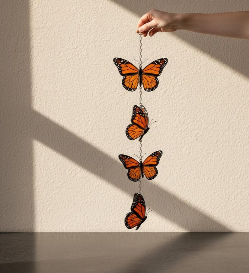 Puede incluir: Colgante decorativo con cuatro adornos de mariposas monarca naranjas y negras suspendidos por una cadena plateada. Las mariposas son sostenidas por una mano sobre un fondo neutro.