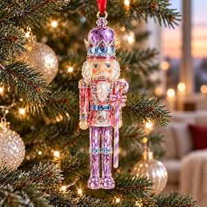 May include: A faceted crystal nutcracker ornament in shades of pink, purple, and blue. The figure wears a crown, has a white beard, and holds a striped candy cane. It is suspended by a red ribbon and features intricate geometric crystal detailing.