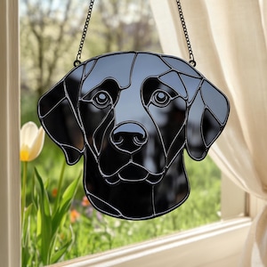 Könnte beinhalten: Schwarze Glasmalerei-Hängedekoration mit dem Kopf eines Labrador Retrievers. Das Gesicht des Hundes ist in geometrische Formen unterteilt, mit einer Kette zum Aufhängen. Der Hintergrund zeigt ein Fenster mit Blick auf einen Garten.