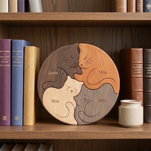 May include: A circular wooden puzzle with four cat-shaped pieces in shades of brown, orange, cream, and grey. The names "Luna", "Milo", "Max", and "Bella" are engraved on each piece. The puzzle is displayed on a wooden shelf.