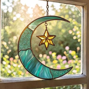 Puede incluir: Media luna de vidriera en tonos turquesa y aguamarina, adornada con un colgante de estrella dorada. La pieza decorativa cuelga de una cadena, perfecta para la decoración de ventanas o paredes. El vidrio tiene una textura.