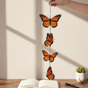 Puede incluir: M&oacute;vil decorativo colgante con cuatro adornos de mariposas monarca naranjas y negras suspendidos por una cadena. Las mariposas se exhiben contra una pared neutra, con un cuaderno y un bol&iacute;grafo sobre una superficie de madera.