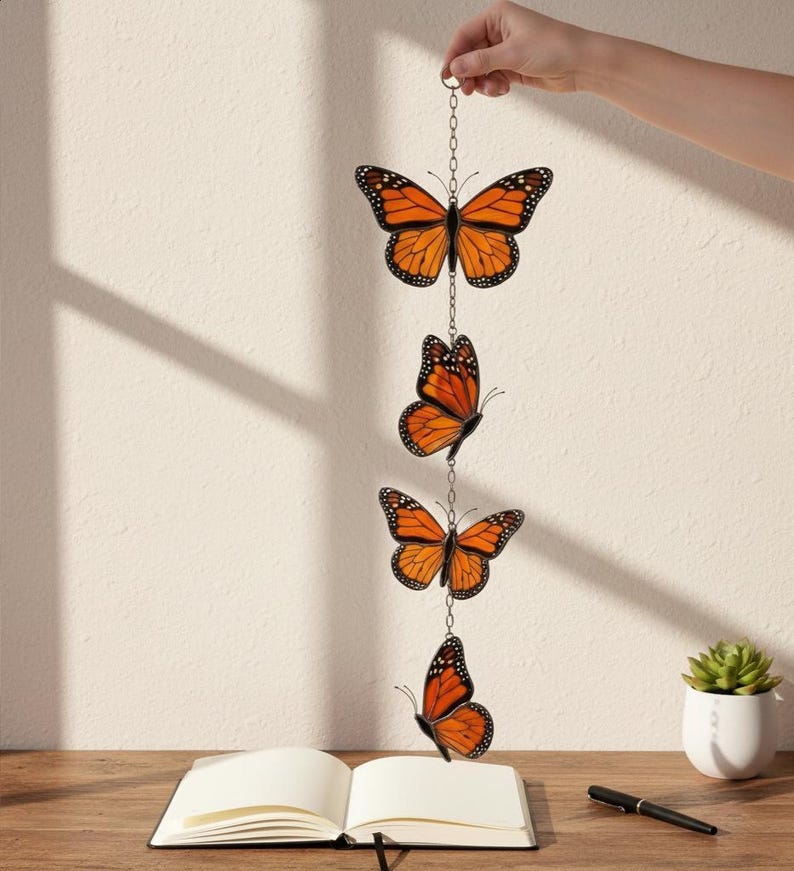 Puede incluir: M&oacute;vil decorativo colgante con cuatro adornos de mariposas monarca naranjas y negras suspendidos por una cadena. Las mariposas se exhiben contra una pared neutra, con un cuaderno y un bol&iacute;grafo sobre una superficie de madera.