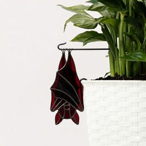 Puede incluir: Una decoración de murciélago de vidriera con alas rojas y negras cuelga de un gancho de metal negro. El murciélago está suspendido frente a una maceta blanca tejida con una planta verde en su interior.