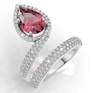 May include: A silver ring featuring a pear-shaped, ruby-coloured gemstone at the centre, encircled by a halo of small, clear stones. The ring's band is split and embellished with more small, clear stones.