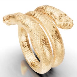 May include: A gold-coloured ring in the shape of a snake. The snake's body coils around the finger, with the head and tail extending outwards. The ring has a textured surface, giving it a realistic appearance.