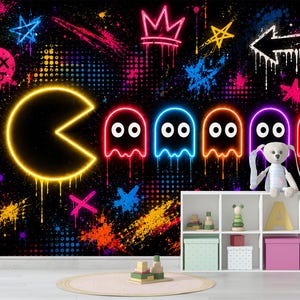 May include: A wall mural featuring a neon-style Pac-Man and five ghosts in red, blue, orange, and purple against a black graffiti background with colorful paint splatters, stars, and a crown, positioned above a white six-cubby storage unit.
