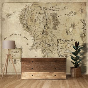 Peut inclure: Une grande carte de la Terre du Milieu est affichée sur un mur, avec les mots "Middle Earth" et "the Sundering Seas". Un meuble de rangement en bois à trois tiroirs est situé en dessous. Un lampadaire et une plante en pot complètent la scène.
