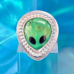 May include: A silver-toned ring featuring a green alien face with black eyes. The alien is set in a teardrop-shaped frame with a beaded border. The ring is displayed on a clear stand against a blue background.