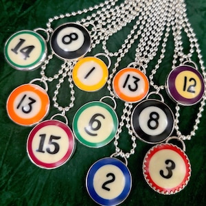 May include: A selection of silver ball chain necklaces, each with a pendant resembling a billiard ball. The pendants show numbers 1, 2, 3, 6, 8, 12, 13, 14, and 15, with different coloured borders.